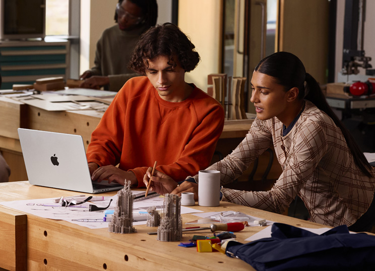 Dos personas colaboran en un proyecto en una mesa, una escribe en un MacBook, la otra usa un lápiz y una regla en papel, hay materiales de arte a su alrededor.