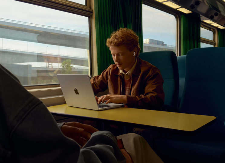 Una persona en el asiento de un tren junto a la ventanilla que usa unos AirPods y trabaja en un MacBook.
