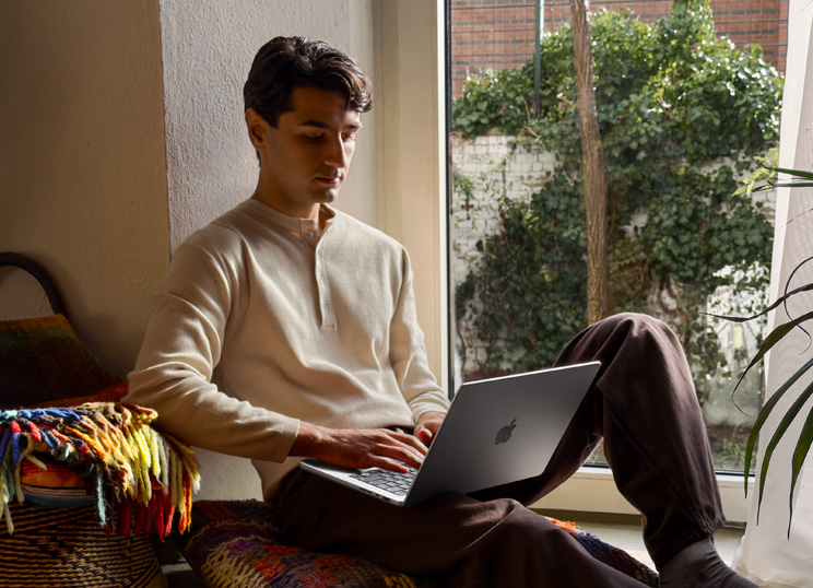 Una persona sentada en el suelo usando un MacBook junto a una ventana.