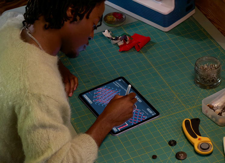 Una persona dibuja una ilustración con el Apple Pencil en un iPad, sentada junto a una base de corte verde con materiales de costura.
